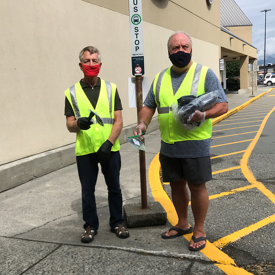 Two men in high visibility vests and masks hold up free masks with tongs at a bus stop.
